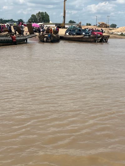 The image shows a busy river crossing scene in Nemnei, Nigeria. Several wooden boats are docked or in the water, some carrying passengers and vehicles. On the larger boat in the foreground, there are at least three cars, including a red sedan and two dark SUVs. Several people are standing on this boat and around it. To the left, another wooden boat is being loaded with cargo and people, with individuals actively involved in the process. In the background, the shoreline is visible, with more people gathered, some under umbrellas. Construction equipment, including a tall crane, is present on the bank, suggesting ongoing development. The river is muddy brown, with visible ripples on the surface. The sky is cloudy.