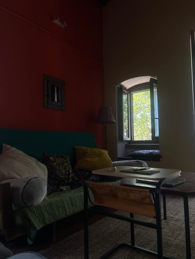 The image depicts a dimly lit interior scene, likely a living area, with a large window offering a view of lush green foliage outside. The room is furnished with a teal-colored couch covered with various pillows, including a patterned floral one and a textured yellow one. A small side table with a light-colored top is positioned in front of the couch, holding a tablet and a pen. To the right of this table, a darker, larger table is visible. A floor lamp with a purple shade stands to the left of the couch, near the window. The walls are painted red on the left and a neutral tone on the right. A dark, recessed wooden frame with a small opening is mounted on the red wall. The flooring appears to be a woven rug. The scene appears to be located in Militello in Val di Catania, Italy, though no specific indicators within the image confirm this location.