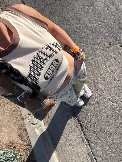 The image shows a person standing on concrete, likely a sidewalk or road, with a partial view of their outfit and surroundings. The person is wearing white sneakers, beige pants with a splatter pattern, and a beige tank top with brown trim and the word "BROOKLYN" printed on it. They have several bracelets on their left wrist and a brown bag with a silver buckle and zipper. Their hair is styled in a braid. The concrete surface shows signs of wear and there is a curb and some ground cover visible in the background. The location is Newport Beach, United States of America.