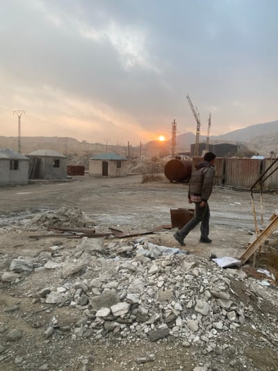 A man in a jacket and dark pants stands amidst a construction site or quarry. Overhead cranes are visible in the distance against a hazy sky with the sun peeking through. The ground is rocky and dusty with piles of debris. Simple concrete buildings are dotted along the landscape. The setting sun casts a warm glow over the scene.