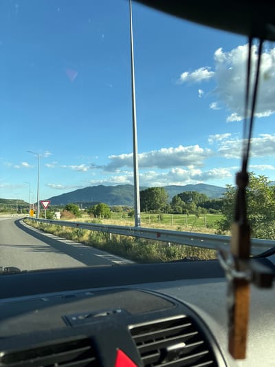 The image presents a roadside view from inside a vehicle in Jeleznitsa, Bulgaria. The vehicle is traveling on a paved road with a metal guardrail on the right side. A mountain range stretches in the background under a partly cloudy sky. Roadside infrastructure is visible, including lamp posts and road signs. The interior of the vehicle, including the dashboard, a wooden cross hanging from the rearview mirror, and air vents, is partially visible.