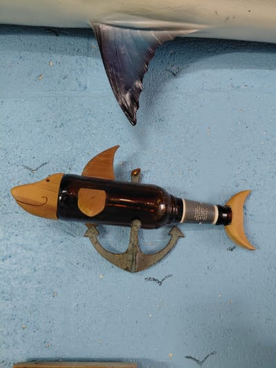 Here is a detailed description of the image:
The image shows a wall decoration in the shape of a fish, composed of a brown beer bottle with wooden attachments. A wooden crescent moon shape is affixed at the top of the bottle, forming the dorsal fin. Wooden pectoral fins are attached to the sides, and a curved wooden piece acts as the tail and lower part of the fish's head, with a small hole representing the eye and a curved line showing the mouth. An old, weathered anchor is attached to the middle of the bottle. To the left of the fish is a partial image of a printed fish fin, and to the right is a partial view of a wooden board. The wall has a textured, light blue finish.
The location is presumed to be Cutler Bay, United States, based on the prompt's instruction, though there are no inherent geographical clues in the image.