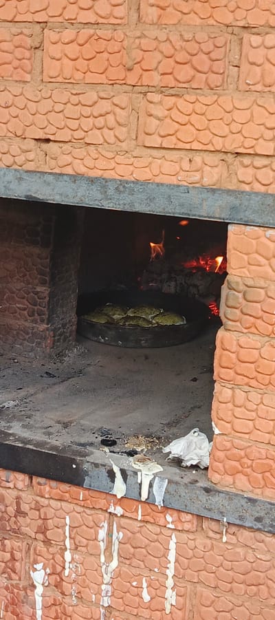 The image shows a close-up view of a rustic, brick oven used for cooking. The oven is constructed with textured, orange bricks that have a pebble-like pattern. Inside the oven, a fire is burning, with visible flames and glowing embers, indicating it's actively in use. In the foreground, a dark, round metal pan is placed on the oven's floor, containing several rounds of what appear to be baked goods, possibly bread or pastries, with a greenish hue. The floor of the oven and the hearth in front of it are dusty and show signs of ash and debris from previous fires. There are also some white paper-like materials, possibly used for cleaning or packing, resting on the hearth. The overall impression is one of a traditional, perhaps rural, cooking setup in Старые Быги, Russia. The lighting suggests it could be daytime, but the interior of the oven is dominated by the warm glow of the fire. There is no visible text in the image.