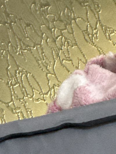 The image is a close-up of a pink fluffy object, likely a stuffed animal or toy, resting on a textured gold surface. A dark cord runs across a grey surface in the foreground. The scene is indoors and there are no discernible features of Ignatievo, Bulgaria present.
