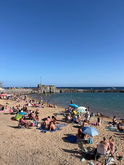 The image shows a beach scene with a clear blue sky. The beach is sandy with a small body of water behind it. There is a large rock formation in the water and a small, white boat is visible in the distance. There are numerous people on the beach, some sitting, some swimming, and some sunbathing. The water appears shallow and clear, with people swimming and wading in the shallows. There are many beach umbrellas scattered on the sand. The scene is located at Playa de Lomo Quiebre in Gran Canaria, Spain. The coordinates 27.818, -15.763 match the location of the beach in the image.