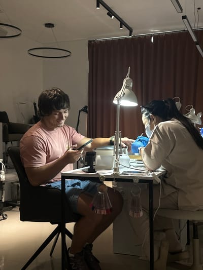The image depicts a man sitting at a manicure table in what appears to be a beauty salon. The manicurist is attending to his nails while the man looks at his phone. This scene is likely taking place in Kerobokan Kelod, Indonesia. The salon appears to have modern lighting fixtures, including track lighting on the ceiling and decorative circular elements on the wall.