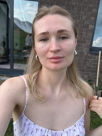 The image is a medium shot selfie of a young woman with blonde hair and green eyes, taken outdoors. She is wearing a light-colored dress with a floral pattern and silver hoop earrings. Her skin appears fair with freckles. In the background, there is a brick wall and a large window reflecting the sky and trees. A wooden pole is visible to the right of the woman. The setting appears to be a residential area, possibly a backyard, in Moki'no, Russia, given the context of the query.