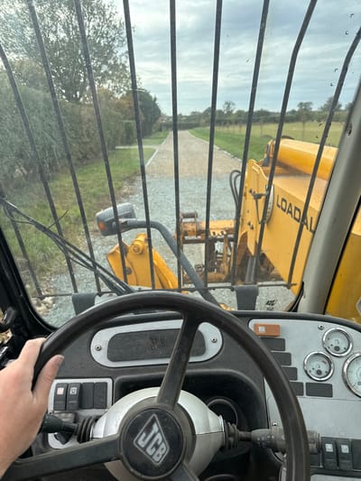 The image depicts the interior of a JCB Loadall telehandler cab, viewed from the operator's perspective, located in Skipsea, United Kingdom. A human left hand rests on the black JCB-branded steering wheel, positioned in the lower center. The dashboard to the right of the steering wheel displays multiple analog gauges for fuel, engine temperature, and RPM, along with various control switches and an orange indicator light. Vertical metal bars of a protective grating are visible through the windshield.
Beyond the windshield, the yellow boom and front mechanism of the JCB Loadall are visible, with the word "LOADALL" stenciled on the side. A light-colored gravel driveway extends into the distance, flanked by manicured green grass verges and hedges. On the left side of the driveway, mature trees and a partial building or structure are visible. The right side of the view shows open ground leading to a line of trees in the background. The sky is uniformly overcast with a grey cloud cover.