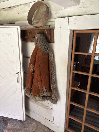 This image shows an interior scene within a rustic, log-built structure in Sielachowskie, Poland. A heavy, brown, fur-lined coat is hanging on a wooden rack mounted on a white-painted wooden wall. To the left, a white-painted wooden door with diagonal paneling is ajar. To the right, a wooden lattice window frames a view into another room, where a patterned wallpaper and furniture are visible. An old wooden sieve or colander is resting on a beam above the coat. The floor appears to be made of stone or concrete. The overall impression is one of an old, well-worn dwelling, possibly a historical or preserved building.
