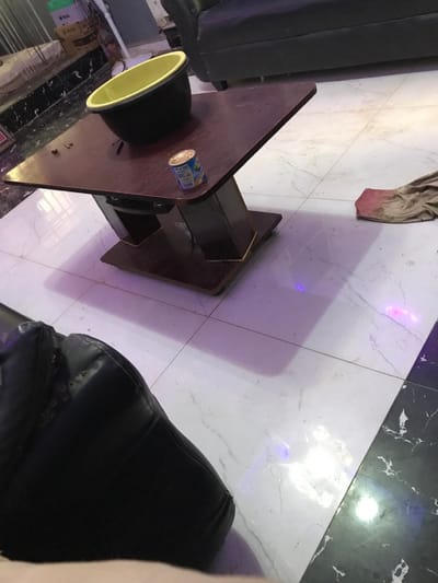 A table with a can and a black object is positioned on a tiled floor with white and black sections. A dirty rag lies nearby. To the left of the table is a black object, possibly a chair. A dark-colored chair or similar object appears to the right. The room is illuminated with an artificial light source causing reflections on the floor.