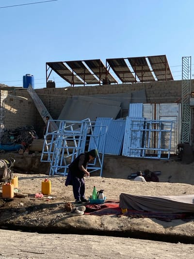 The image portrays an outdoor commercial or workshop area in Kōkrān, Afghanistan, under a clear blue sky. In the foreground, a person wearing a dark vest, blue tunic, and dark cap stands, bent over a blue plastic basin containing a green bottle, a clear bottle, and a black thermos. A pair of light-colored shoes rests on the dry, dusty ground near their feet. A red patterned rug is spread on the ground to their right, partially covered by a light-colored fabric. Further to the right and slightly lower, two additional individuals are seated on the ground, engaged in an activity.
The midground features various materials and structures: a large quantity of stacked light-colored metal frames, possibly window or door frames, and multiple metal sheets and panels, some with decorative ironwork, leaning against a wall. To the far right, another person is visible working near metal structures. On the left midground, a pile of scrap metal, including bicycle frames and tires, is present, alongside a partially visible blue industrial machine or compressor.
The background consists of a rough brick wall. On the rooftop behind this wall, two distinct arrays of solar panels are mounted on metal frames. To the far left, a blue plastic water tank is visible on a raised platform. A large, angled sign, unreadable, leans against the wall, and piles of corrugated metal sheets are present.