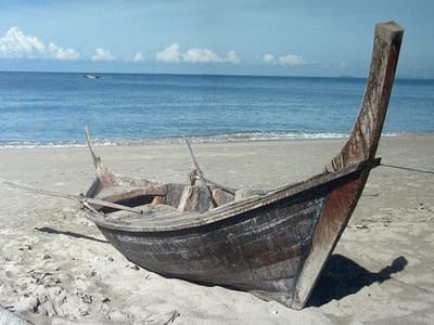 A weathered, wooden fishing boat is beached on a sandy shore. The boat is dark brown and appears old, with visible planks and signs of wear and tear. A long, wooden oar is propped against the side of the boat, extending outwards. The boat is angled, with its bow pointing towards the right of the frame and its stern towards the left.
The setting is a beach with a gentle slope leading down to the ocean. The sand is a light tan color and appears dry, with some scattered debris. The ocean is a calm, deep blue, with small waves lapping at the shore. In the distance, the horizon is a clear line between the sea and the sky. The sky is a pale blue, with a few scattered white clouds. There is another, smaller boat visible in the far distance on the water, which appears to be moving. The overall impression is one of a quiet, possibly isolated, coastal scene. The lighting suggests it is daytime, likely late morning or early afternoon, with a bright sun casting shadows. There is no visible text in the image.