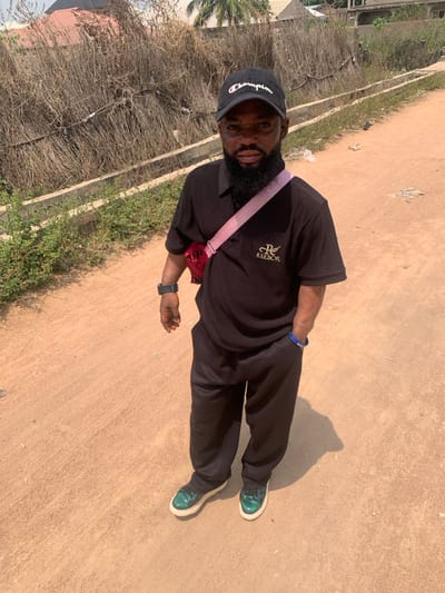 An adult male stands on an unpaved, light brown dirt path in Eika-Adagu New Layout, Nigeria, facing the camera directly. He wears a black ribbed short-sleeved polo shirt with a gold-colored "REEBOK" logo on the left chest, loose black trousers, and dark green/teal metallic-sheen sneakers with white soles. A black baseball cap with a white "Champion" logo is on his head. He has a full dark beard. A red fanny pack with a pink strap is worn crossbody over his right shoulder and across his chest. A black digital watch is on his left wrist, and a blue wristband is on his right wrist. The path shows minor debris and shadows from the subject. In the immediate background to the subject's left, a low concrete barrier separates the path from an area with sparse green vegetation and a dense mass of dry, brown, fibrous vegetation. Distant residential structures with light-colored walls and dark roofs are visible further in the background under a clear, light blue sky.