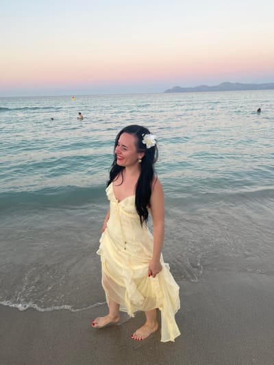 A woman stands on a beach in Platja De Muro, Spain, at dusk, looking out at the ocean. She has long black hair adorned with a white flower and is wearing a light yellow dress. The ocean is calm with small waves gently lapping at the shore. In the distance, there are a few people swimming and a red and yellow float. The sky is a gradient of pink and purple, indicating the sunset. The sand is a light gray color. The overall scene evokes a peaceful and serene atmosphere.