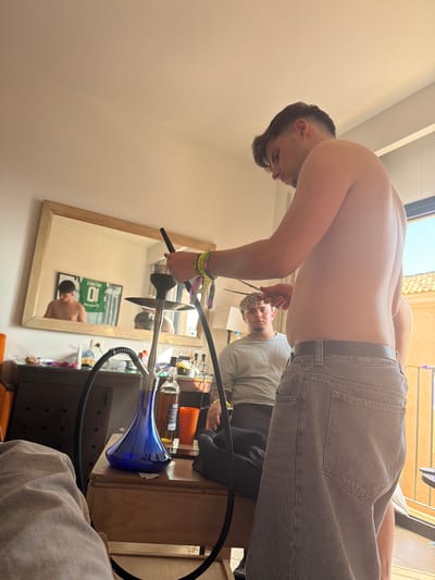 A slightly rotated medium shot focuses on a shirtless man with dark hair, wearing gray jeans, setting up a hookah. He is leaning over a table with a mirror. The reflection in the mirror shows him as well as another man in a white shirt sitting down, looking at a phone. Also in the mirror is a soccer jersey on the wall. A blue hookah base is visible on the table, along with various liquor bottles and accessories. The environment appears to be inside a room, likely a residence, in Punta Umbría, Spain.