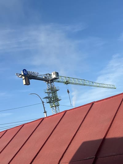 This is a low-angle, full-shot photograph of a construction crane against a bright blue sky with scattered white clouds. The crane is predominantly green and white and features a large, blue circular logo with white lettering that reads "AI GROUP" on its horizontal jib. A white operator's cabin is visible near the center of the crane. Below the crane, a section of a red, metal-seamed roof occupies the foreground. A street light is visible to the left of the crane's tower. The scene is set in the city of Lebedinovka, Kyrgyzstan.