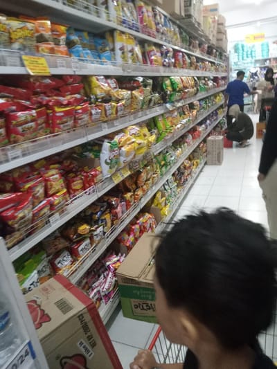 This image depicts an interior view of a supermarket or grocery store aisle in Nganjuk, Indonesia. The primary focus is a long, brightly lit aisle extending into the background, lined on the left by white metal shelving units fully stocked with numerous packaged snack foods, including chips, crackers, and instant noodles, in colorful plastic and foil packaging. Yellow promotional labels are affixed to some shelf edges.
In the foreground, the back of a person's head with short, dark hair, likely a child, is visible on the right, looking towards the right side of the aisle. Their hands rest on the red handle of a partially visible shopping cart. To the left of this person, a cardboard box for "Happy Tie Tic" brand snacks is on the floor, next to a smaller, unidentified cardboard box. On the far left, a clear plastic water bottle and a brown cardboard box with red branding and "NET. 2000 PCS" text are partially visible on a lower shelf.
Further down the aisle, towards the midground on the right, a person wearing a dark olive-green top and a light-colored head covering is crouching down, appearing to stock or inspect items on a lower shelf. Behind them, another individual in a blue shirt stands upright, facing away from the camera, near another section of the store. A third person is partially visible on the far right. The floor is covered with white tiles, and the overhead lighting is bright, consistent with a retail environment. Several cardboard boxes are stacked on the floor near the crouching individual.