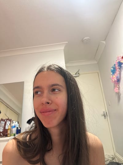 The image displays a young female subject indoors, located in Corby, United Kingdom. The subject, positioned slightly to the right of the frame's center, possesses long, straight, dark brown hair and is looking towards the upper left with a subtle smile. Her right shoulder is bare. The room features white walls and ceiling with white crown molding. A white smoke detector is affixed to the ceiling in the upper right quadrant. A light-colored door with a silver handle is visible to the right. A decorative pink and blue tinsel unicorn figure is mounted on the wall to the right of the door. The left background shows a partial mirror reflection of various bottles and a brown package on a surface. Some undefined clutter is visible on the floor in the lower right.