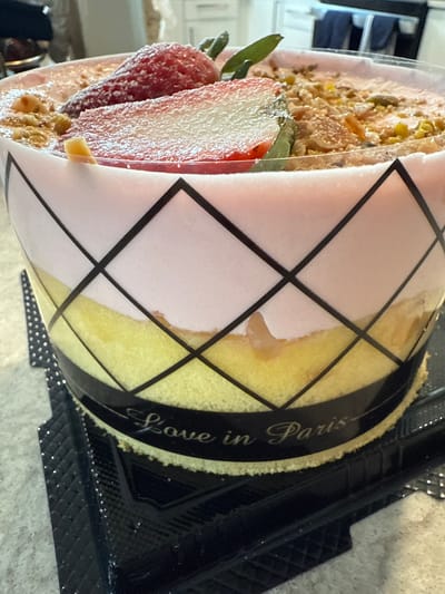 A slice of cake in a transparent plastic container with a black rim sits on a neutral surface. The cake has a pink layer and a yellow layer, visible through the container's graphic design of intersecting black lines. The label on the container reads "Love in Paris". The cake is topped with a sliced strawberry and a crumbly topping. A black plastic tray with textured edges is partially visible beneath the container. The location of the image is the city of Orlando, United States.