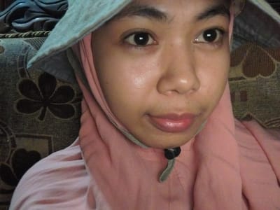 This is a close-up, medium-shot photograph taken outdoors in Labissa, Indonesia. The subject is a young person, likely female, wearing a pink hijab and a light blue sun hat. The person is looking directly at the camera with a neutral expression. The background consists of a patterned sofa. The lighting appears to be natural daylight, suggesting it might be daytime. There is no visible text in the image.