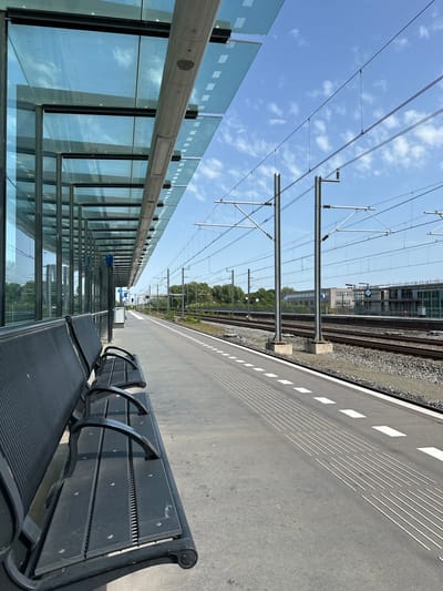 The image shows a train platform in Dronten, Netherlands, with benches in the foreground. A glass wall runs parallel to the platform edge. Railway tracks are visible on the right, alongside a building. Overhead power lines stretch above the platform, and a blue sky with scattered clouds is in the background.