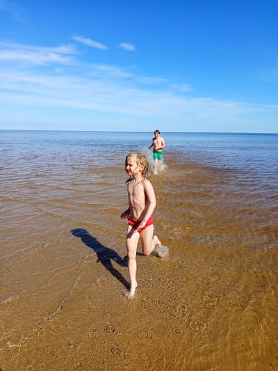 The image depicts two children running through the shallow waters of a large body of water, likely a lake or wide river, near Podborov'e, Russia. The scene unfolds under a bright, clear blue sky with minimal, wispy white clouds.

In the foreground, a young girl, estimated to be between 4 and 7 years old, with light-colored, wet hair, runs barefoot towards the right side of the frame. She is wearing red swim briefs and is bare-chested. Her right foot is submerged, actively splashing water, while her left foot is lifted in mid-stride. Her head is turned slightly to her right, and her mouth is partially open, indicating exertion or excitement. A distinct, elongated shadow of her body is cast to her left on the sandy bottom.

Further back and to the right, an older boy, estimated to be between 8 and 12 years old, with short, dark hair, runs behind the girl. He is wearing green swim trunks and is also bare-chested. He is mid-stride, producing larger splashes of water around his legs, suggesting he is moving faster or with more force.

The water near the shore is very shallow and clear, revealing a light brown, sandy or muddy bottom with visible ripples and concentric splash patterns created by the children's movement. As the water extends towards the horizon, it gradually deepens and transitions to a darker blue hue, meeting a flat horizon line. The strong sunlight casts clear shadows and illuminates the water, making it appear golden-brown near the foreground. Both children are actively engaged in running and splashing, indicating play.