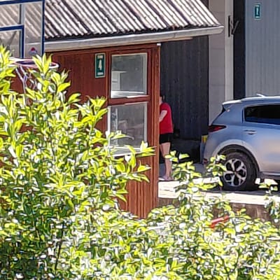 In Skuķīši, Latvia, a person in a red shirt and dark shorts lies on their side, appearing to be resting or possibly sleeping. They are positioned on a raised platform or ledge, which is a dark, possibly wooden surface. In the background, a silver car is parked, with its wheel clearly visible. The scene is framed by lush green foliage in the foreground and to the right, suggesting an outdoor setting with trees or bushes. To the left of the person, there is a dark-colored building or structure with a window. Below the window, a rectangular green sign with a white symbol is visible; the symbol appears to be an arrow pointing to the right within a horizontal bar. The lighting suggests it is daytime, with sunlight casting shadows and highlighting the leaves. The overall mood is relaxed and tranquil.