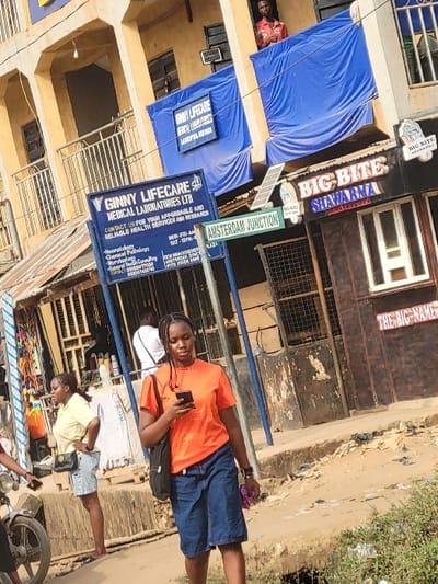 The scene depicts a daytime street view in Puerto López, Colombia, at a location identified by a street sign as "AMSTERDAM JUNCTION." In the foreground, a young adult female with dark braided hair walks towards the viewer's right, looking down at a black smartphone held in both hands. She wears an orange short-sleeved shirt, dark blue denim shorts or skirt, and black shoes, carrying a black shoulder bag. Wired earbuds are visible connected to her phone. To her immediate left, partially obscured by the front wheel and engine of a light-colored motorcycle, another young adult female stands with her hands on her hips, wearing a light yellow collared shirt and light blue denim shorts, with a black shoulder bag.
The background features multi-story buildings with light brown/beige facades, some displaying wear and exposed construction. A prominent blue and white sign for "GINNY LIFECARE MEDICAL LABORATORIES LTD" is mounted on a pole, detailing services such as Haematology and Microbiology. A smaller blue banner for "GINNY LIFECARE" hangs from a balcony above. Adjacent to these, a green street sign reads "AMSTERDAM JUNCTION," with a smaller sign beneath it partially legible as "PLAZA." Above the junction sign, a small solar panel is affixed to a pole. To the right of the central focus, a commercial establishment named "BIG BITE SHAWAPMA" is visible with a dark brown sign featuring white and red lettering and yellow backlighting. Another sign, "THE BIG-NAME," is partially seen further right. Blue tarpaulins are draped over balconies on the upper floors of the background buildings. From a window or balcony on an upper floor, a young adult male in a red patterned shirt observes the street below. The ground is unpaved, appearing as dirt or mud with visible debris and uneven terrain.