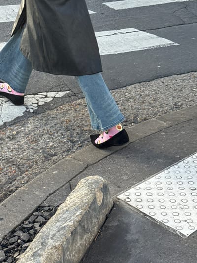 The image displays the lower portion of an individual walking on a public street, presumably in Asnières-sur-Seine, France; the specific location within the city is not visually identifiable. The person is wearing a long, dark-colored coat, light blue denim jeans, pink socks adorned with a repetitive pattern of green avocado halves, and black pointed-toe shoes with a wedge heel. The subject is captured mid-stride, with their right foot on the asphalt pavement near a concrete curb. A white-striped pedestrian crosswalk is visible on the dark grey asphalt road surface. The environment includes a rough-hewn, light grey stone block partially in the foreground on the sidewalk, adjacent to a rectangular silver metallic tactile paving plate featuring raised circular studs. Small aggregate debris is present in the gutter alongside the curb. Natural daylight illuminates the scene.