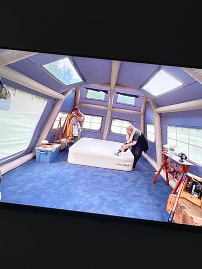 In Rosenovo, Bulgaria, a large inflatable tent, likely a portable dwelling or event space, is depicted. The interior is carpeted in blue and features several windows and skylights that allow natural light to enter. A white air mattress, labeled "RestBed," is centrally placed. A woman with blonde hair is vacuuming the mattress. To the left, a man is standing next to a wooden rack with several garments hanging on it. A blue storage bin and a small lamp are on a side table. To the right, a wooden table holds a plant and a digital clock displaying "13:38." Below the table is a wooden chest containing various items. The overall scene suggests preparations for occupancy or an event within the temporary structure.