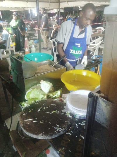 The image captures an outdoor or semi-sheltered food stall, likely operating at night or in low-light conditions, illuminated by artificial overhead lighting. The scene is consistent with an informal street food establishment in an urban area like Makadara/Mji Wa Kale ward, Kenya.
In the immediate foreground, a dark, circular wooden chopping board is present, covered with white shredded cabbage and other green vegetable scraps. To its left are larger, fresh pieces of white cabbage. Adjacent to the chopping board, a stainless steel deep fryer is visible, containing a rectangular food item immersed in hot oil, with a pair of metal tongs resting on its edge. To the right of the deep fryer, a large, empty turquoise plastic basin is positioned.
A dark-skinned male chef, wearing a grey short-sleeved polo shirt, a blue apron bearing the text "Unga ni TAIFA" (Flour is Nation) with a map of Kenya logo, and white disposable gloves, stands centrally, actively engaged in food preparation. His right hand holds a utensil, possibly tongs, over a cooking area. His left hand is positioned near a gas burner connected to a blue gas cylinder visible below the counter. To the chef's right, a yellow plastic bowl holds a liquid, likely oil or batter. Below this, a stack of white, disposable paper plates, encased in clear plastic, rests on the counter. A tall, clear plastic beverage dispenser, filled with an orange liquid, is partially visible on the far right.
In the midground and background, under a light-colored tent canopy supported by metal poles, several other individuals are present. To the far left, a person in a light blue shirt is partially visible, seated at a white plastic chair. To the left of the deep fryer, a woman dressed in a black garment, a light green headscarf (hijab), and carrying a brown shoulder bag, stands, observing the activity. Further back, a man in a dark t-shirt and light-colored pants stands. To the right, a woman with short dark hair, wearing a dark top, is seated at a white plastic chair. Multiple white plastic tables and chairs are scattered throughout the background, indicating a dining area. A red SUV is partially visible in the distant background, suggesting proximity to a street or parking zone.