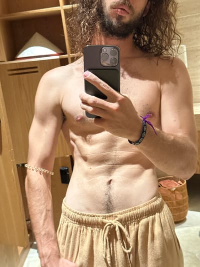 The image displays a mirror selfie of a person, likely male, with long, curly dark hair and a beard, holding a dark smartphone, possibly an iPhone, to capture the reflection. The person is shirtless, revealing a light-skinned torso with visible body hair on the chest and abdomen. They are wearing light brown or tan drawstring shorts with an elasticated waistband.
On the left wrist, two bracelets are visible: a thin purple string bracelet tied with a bow, and a dark blue patterned bracelet with white diamond shapes below it. A gold-colored beaded bracelet is worn on the right wrist.
The setting appears to be indoors, likely a room or bathroom, with warm, natural light casting distinct shadows. Partially visible in the background are a wooden shelving unit on the left and a woven basket on the right. The image is presented rotated 90 degrees counter-clockwise. No visible text is present. The provided context indicates the location is Mu Cang Chai, Vietnam, suggesting this is an interior scene within that region. The overall impression is casual.