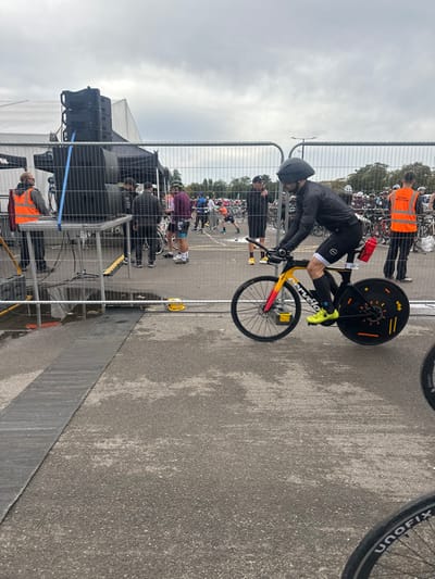 The image depicts an outdoor event, likely a triathlon or cycling race, situated in Weymouth, United Kingdom. The ground consists of wet, dark asphalt pavement, indicating recent precipitation, with a dark rubber mat or cable protector running along the left side. The sky is uniformly overcast.
In the left midground, a large, temporary sound system is positioned behind a metal crowd barrier, consisting of stacked black speaker cabinets on a raised platform, adjacent to a black event tent. A male individual wearing an orange high-visibility vest stands near this equipment. Puddles are visible on the asphalt behind the barrier.
The central subject is a male cyclist mounted on a high-performance Cervélo time trial bicycle. The cyclist wears a black aerodynamic helmet, a black long-sleeved cycling top, black cycling shorts with a white stripe on the left thigh and a red race number "231" on the right thigh, and bright yellow cycling shoes. His hands are positioned on the bicycle's aerobars. The bicycle features a distinctive yellow-to-red gradient paint scheme on the front fork and main frame, a black spoked front wheel with a disc brake, and a solid black disc wheel with orange markings on the rear. A red water bottle is attached behind the seat.
In the background, beyond additional metal barriers, numerous individuals are present, many dressed in cycling attire, alongside a multitude of bicycles racked in what appears to be an event transition or bike parking area. A second individual in an orange high-visibility vest is visible to the right of the central cyclist. A large white event tent and distant foliage are discernible under the cloudy sky. A partial view of another bicycle wheel with "UNOFX" text is in the bottom right corner of the frame.
