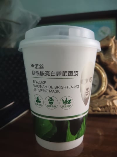 The image shows a white cylindrical container with a white lid, consistent with cosmetic packaging. The container has text and graphics printed on it. The text in Chinese appears to be "希诺丝 烟酰胺亮白睡眠面膜," which translates to "Xinuo Si Niacinamide Brightening Sleeping Mask." Below this, in English, it reads "SEALUXE NIACINAMIDE BRIGHTENING SLEEPING MASK." There are three circular icons below the English text. The leftmost icon depicts a crescent moon and stars. The middle icon shows a face with dark circles under the eyes. The rightmost icon features a stylized plant with leaves. To the right of the text, there is an embossed image of a human face. The lower part of the container features a green and black design of large leaves. The container is placed on a dark wooden surface. In the background, a blurred object with a golden metallic frame, possibly a clock or a decorative item, is visible. The lighting suggests an indoor setting. There is no information in the image that would indicate its location in Sharg'un, Uzbekistan, or any specific urban environment.
