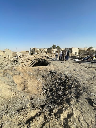 The image depicts a scene of extensive structural damage in Spin Boldak, Afghanistan, under a clear, cloudless blue sky.
In the immediate foreground, a significant, roughly circular crater is present on the uneven, dusty ground, approximately 2-3 meters in diameter, surrounded by excavated earth and various fragments of rubble. The ground surface consists of compacted soil, gravel, and scattered debris. Shadows are cast towards the lower right, indicating a high sun angle.
Midground features a group of six individuals, primarily adults, standing on a relatively clear patch of ground amidst the destruction. They are dressed in traditional light-colored garments, including long shirts, trousers, and head coverings. The individuals appear to be observing the surrounding devastated area; no active engagement with the debris is visible.
Behind the individuals, an expansive area of severely damaged and collapsed buildings extends across the midground and into the background. Structures are reduced to piles of rubble, partially standing walls constructed from what appears to be mudbrick or concrete, and exposed skeletal remains of roofs and wooden beams. The widespread destruction is consistent with a catastrophic event such as a large-scale explosion. Scattered debris, including bricks, concrete blocks, and miscellaneous building materials, covers the entire scene. A few small, green trees are visible in the distant background, providing a slight contrast to the otherwise desolate, demolished environment.