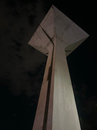This media file is a nighttime, low-angle shot of a concrete structure, likely a monument or a piece of modern architecture, set against a dark sky with scattered clouds. The structure is angular and abstract, with sharp edges and smooth, curved surfaces. It appears to be illuminated from below, casting shadows and highlighting its geometric form. A thin, dark line runs along a crevice in the concrete, possibly a joint or a decorative element.
The scene is devoid of people or any overt activity, focusing solely on the imposing presence of the concrete structure. The dark sky suggests it is late evening or night, and the presence of clouds indicates a potentially overcast or dynamic weather condition, though no precipitation is visible. There are faint hints of greenery at the very top right of the frame, suggesting the presence of trees or other vegetation surrounding the structure. A tiny, bright speck in the dark sky, visible in the bottom right quadrant, could be a distant star or a small aircraft light.
No text is visible within the frame. The overall mood is one of quiet grandeur and a sense of mystery, emphasizing the scale and form of the architectural element in the darkness. The location context of Tha Khlong Town Municipality in Thailand is noted, but no specific visual cues within the image definitively confirm this location.