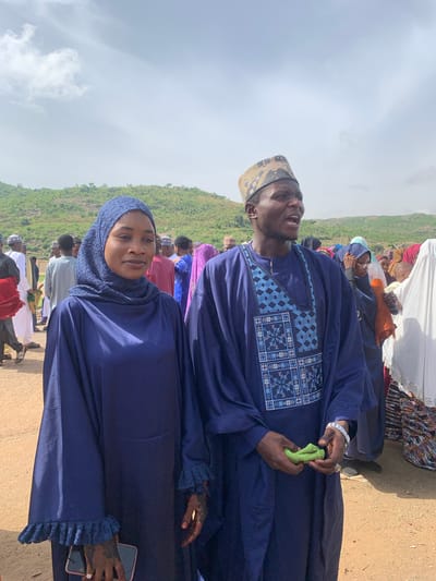 In Naraguta Hausawa, Nigeria, a man and a woman are prominently featured in an outdoor gathering. The man, wearing a blue traditional robe with intricate embroidery on the chest, also sports a patterned cap and a silver watch. He holds a small green object in his hands. Beside him, the woman is dressed in a matching blue hijab and robe, adorned with ruffled cuffs. She holds a mobile phone in her right hand, and both hands display henna designs. They are part of a larger group of people, visible in the background, some wearing hijabs and traditional attire. The terrain appears to be a dirt ground with a lush green hill in the distance under a cloudy sky.