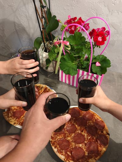 The image depicts a table setting with three glasses of dark liquid, likely soda or juice, and two pepperonis pizzas. Several hands are visible, holding the glasses in a toast gesture above the pizzas. In the background, a potted plant with red flowers and green leaves sits in a pink and white striped bag, behind a textured gray wall. There are no discernible elements that would indicate a specific location within the city of Brodi, Latvia. The scene appears to be an indoor gathering for a meal.