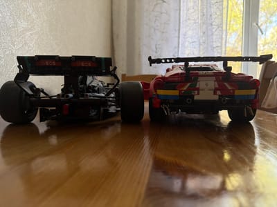 The image shows two LEGO Technic model cars from the rear, standing on a varnished wooden surface, likely a table. The car on the left is black and has "INEOS" printed on its rear wing. The car on the right is primarily red and has "AFCorsa.it" printed on it. A window with thin white curtains is visible in the background. The image does not offer evidence to corroborate the location in Pavlovo, Russia.
