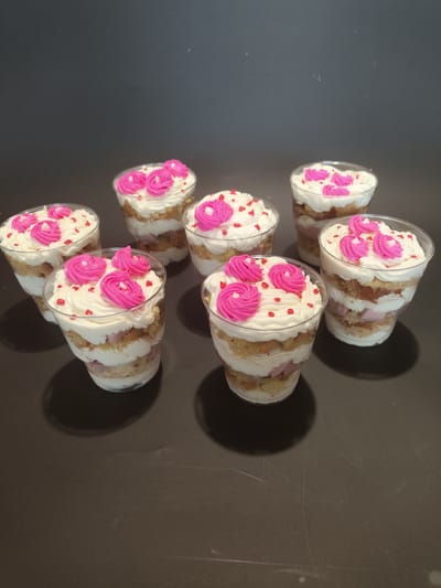 Seven individual servings of trifle are arranged on a dark surface against a dark background. Each trifle is presented in a clear plastic cup and consists of layers of cake, white cream, and pink filling. The trifles are topped with white whipped cream, accented with piped pink frosting in a floral pattern, and sprinkled with small red edible decorations. The lighting casts shadows to the right of the trifles. The image appears to be a studio shot, with no identifiable features of Myadzyel, Belarus.