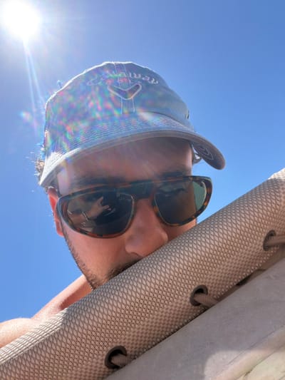 A low-angle perspective shows a person wearing a hat and sunglasses against a bright blue sky. The sun flares intensely to the lower left. Part of a light-colored, textured awning or cover is visible on the right, featuring grommets. The person's face is angled down, with facial hair evident. The hat is light blue with a darker brim, and the sunglasses have a tortoise-shell pattern. A reflection is visible in the sunglasses lenses.