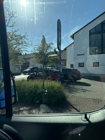 The image captures a street scene in Volpertshausen, Germany, on a bright, sunny day. The perspective is from inside a vehicle, looking out through the windshield and side window. In the foreground, the interior of the vehicle is visible, including a dark-colored dashboard and a portion of the driver's side door.
Outside the vehicle, a paved street or sidewalk with a brick pattern is visible. Several cars are parked or driving on the street. A dark-colored SUV is parked in front of a white building with a red-tiled roof. To the left of the SUV, a red hatchback is also parked. Further down the street, other vehicles and buildings can be seen.
A prominent bus stop sign with a blue pole stands near the parked cars. A yellow diamond-shaped road sign is also visible, mounted on a pole behind a cluster of green bushes. The sun is shining brightly, casting shadows on the ground and creating lens flare in the image, particularly in the upper left quadrant. Trees with green foliage are present on the sides of the street, and the sky is a clear blue. The white building in the background features large windows with blinds and the number "114" is visible on its facade.