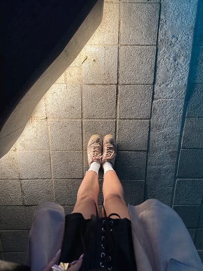 A medium-angle, high-angle shot shows a person's legs and feet standing on a paved surface. The person is wearing light-colored sneakers and white socks. They are also wearing dark shorts with a laced-up detail. Part of a light-colored sweatshirt is visible around the waist. A portion of a dark, angled structure is on the left. The paved surface consists of textured, gray tiles with light reflecting off it. The setting is suggestive of an urban environment. Without additional information, it is not possible to confirm that the location is specifically Buk-myeon, South Korea.