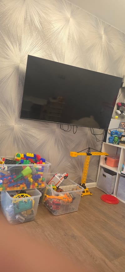 A television is mounted on a wall with a geometric pattern. Below the television, there are several clear plastic bins filled with toys, including building blocks, toy vehicles, and construction toys. A large yellow toy crane stands to the right of the bins. A white shelving unit is also visible to the right of the crane, containing more toys and storage boxes. The flooring is a wood-effect laminate. The image does not provide any information about the location in the city of Berdsk, Russia.