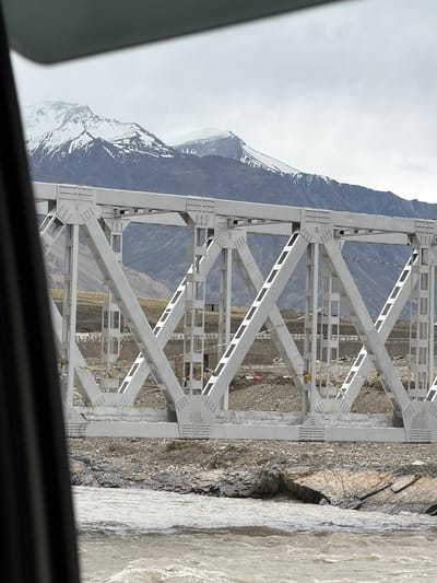 The image shows a white truss bridge spanning a rocky riverbed in Zangla, India. Snow-capped mountains form the backdrop, with a cloudy sky overhead. The bridge is constructed from metal beams and rivets, with diagonal supports creating a geometric pattern. Prayer flags are strung along the bridge, fluttering in the wind. The river below is murky brown and appears to be flowing rapidly. The foreground is dominated by the river and the rocky shore, while the bridge and mountains are visible beyond. The perspective suggests the photo was taken from a vehicle.