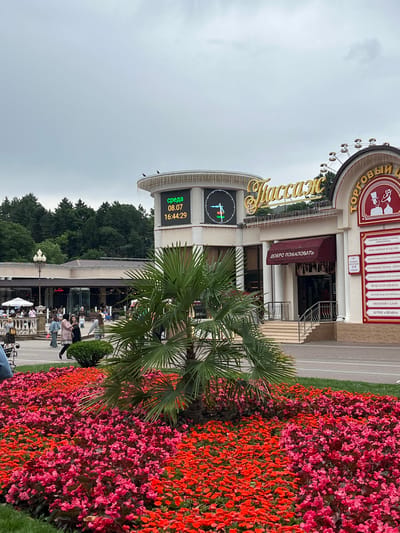 This media file captures a vibrant scene in Kislovodsk, Russia. The foreground is dominated by an abundant flower bed bursting with bright red and pink blooms, interspersed with lush green palm fronds. Behind the flowers, a bustling pedestrian area unfolds.
The main subject is a commercial building with a prominent sign featuring the Cyrillic word "ТОРГОВЫЙ" (meaning "trade" or "commercial") and "ОЛИМПИЕЦ" (Olimpiets, likely a name). Below this, in stylized gold lettering, is the word "Массаж" (Massage). A digital display on the building shows "среда 08.07 16:44:29", indicating it is Wednesday, July 8th, at 4:44:29 PM. This suggests the time of day is late afternoon.
Various shops are listed on the building's facade, including "СПОРТИВНАЯ ОДЕЖДА И ОБУВЬ" (Sportswear and Footwear), "ЮВЕЛИРНЫЕ ИЗДЕЛИЯ" (Jewelry), "ГОЛОВНЫЕ УБОРЫ, ШУБЫ" (Hats, Furs), "МУЖСКАЯ ОДЕЖДА" (Men's Clothing), and "КОРЕЙСКАЯ КОСМЕТИКА" (Korean Cosmetics). A sign above an entrance reads "ДОБРО ПОЖАЛОВАТЬ" (Welcome).
Several people are visible in the background, engaged in various activities. Some are walking, while others appear to be seated at outdoor cafes or browsing shops. The overall atmosphere seems lively and engaging, suggesting a pleasant, possibly warm, day. The cloudy sky hints at overcast weather. The combination of the flowers, the commercial activity, and the presence of people creates a sense of a popular public space.