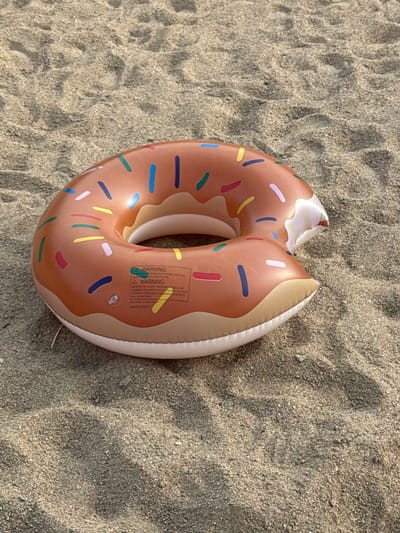 An inflatable donut-shaped pool float lies on a sandy surface. The float is a light brown color with multicolored sprinkles scattered across its surface, and a white frosting detail on one edge. A warning label is visible on the inner ring of the float. The sandy surface appears to be the beach. The location is the village of Volodarsky, Volodarsky rural district, West Kazakhstan Region, Kazakhstan.