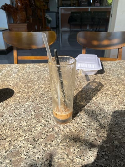 The scene depicts a granite-patterned tabletop in an establishment located in Đak Đoa, Vietnam. In the foreground, a tall, clear glass, approximately two-thirds empty, contains light brown liquid residue at its base. Two straws are inserted into the glass: one clear plastic and one black plastic. To the right, a white Styrofoam takeout container with a lid is present, with two light-colored wooden chopsticks resting horizontally across its top. Strong sunlight illuminates the table, casting distinct shadows from the glass and container. The background, out of focus, reveals two wooden chairs with curved backrests and a large, ornate wooden carving to the left. The floor behind the chairs appears dark and reflective. No individuals are visible.
