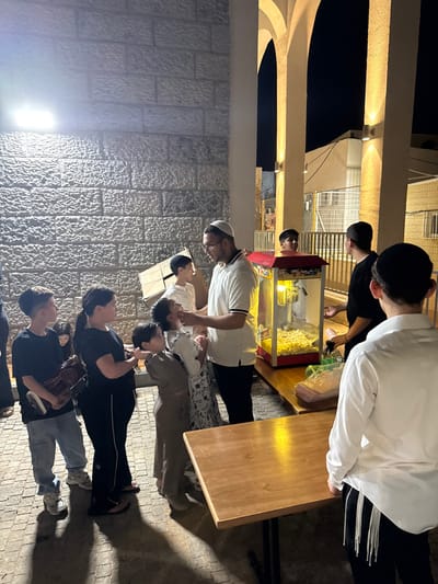 The image captures an outdoor night scene in Beer Yaaqov, Israel, illuminated by artificial lighting. A group of approximately ten individuals, predominantly children and young males, are gathered near a light-colored, textured stone block wall on the left and a series of warmly lit arched columns on the right.

In the central area, a male adult wearing a white polo shirt, glasses, and a white kippah is observed bending down, engaging with a young girl in a patterned dress, appearing to hand her an item. To his immediate left, a young boy in a brown outfit stands. Further left, a line of children includes a girl in a black t-shirt and dark pants, and a boy in a black t-shirt and light blue jeans holding a brown bag. Directly behind the adult male, a boy in a white t-shirt and dark kippah stands near a red and clear popcorn machine, which is full of popcorn and positioned on a wooden table. Several green bags are also on this table. To the right, another wooden table is visible in the foreground, and a young male in a white long-sleeved shirt with visible tzitzit is seen from the back, facing the central activity. Additional children are partially visible in the background and within the group.

The ground is paved with interlocking pavers, and strong shadows are cast by the individuals and objects. A bright floodlight illuminates the stone wall on the left, while the arched columns on the right are highlighted by embedded lighting. Beyond the columns, the dark night sky, portions of a building, and a fence are discernible.