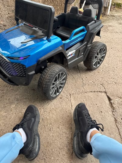 The image presents an outdoor scene in Konyavo, Bulgaria, captured from a slightly elevated perspective looking down. It features a person's lower legs and feet alongside a blue and black toy electric ride-on car, which is tipped onto its side.
The person is wearing light blue sweatpants, white ankle socks, and black athletic sneakers. The sneakers prominently display the Nike brand logo and "AIR MAX" text on their sides, identifying them as Nike Air Max footwear. One foot is positioned closer to the toy car, while the other is slightly withdrawn, indicating an interaction with the vehicle.
The toy car, designed to resemble a small SUV or truck, is predominantly blue at the front and sides, contrasted with black components for its chassis, seats, and grille. It rests on its side, revealing its undercarriage and two of its wheels, which have dirtied tires consistent with outdoor play. A "BMW M" logo sticker is visible on the black rear section of the car's body, along with partially visible text "PER" from what appears to be "PERFORMANCE." Notable details on the car include a patterned front grille, side mirrors, and a blue handle integrated into its design.
The ground is a worn, cracked concrete or paved surface, showing signs of age and use. In the background, glimpses of dirt and minimal vegetation suggest a less manicured outdoor environment. The lighting is bright and even, indicative of daytime.